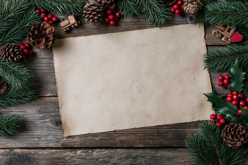 Blank aged paper sheet on wooden surface framed by pine branches, holly, berries and pinecones for christmas decoration