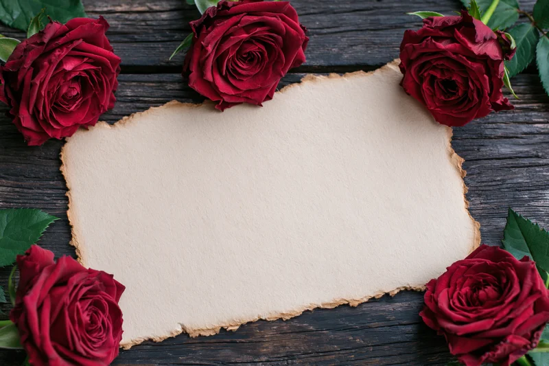 Aged parchment card with torn edges surrounded by red roses on dark wooden surface