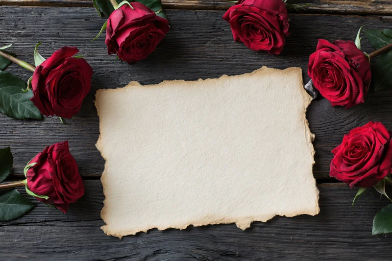 Aged parchment card with torn edges surrounded by red roses on dark wooden surface