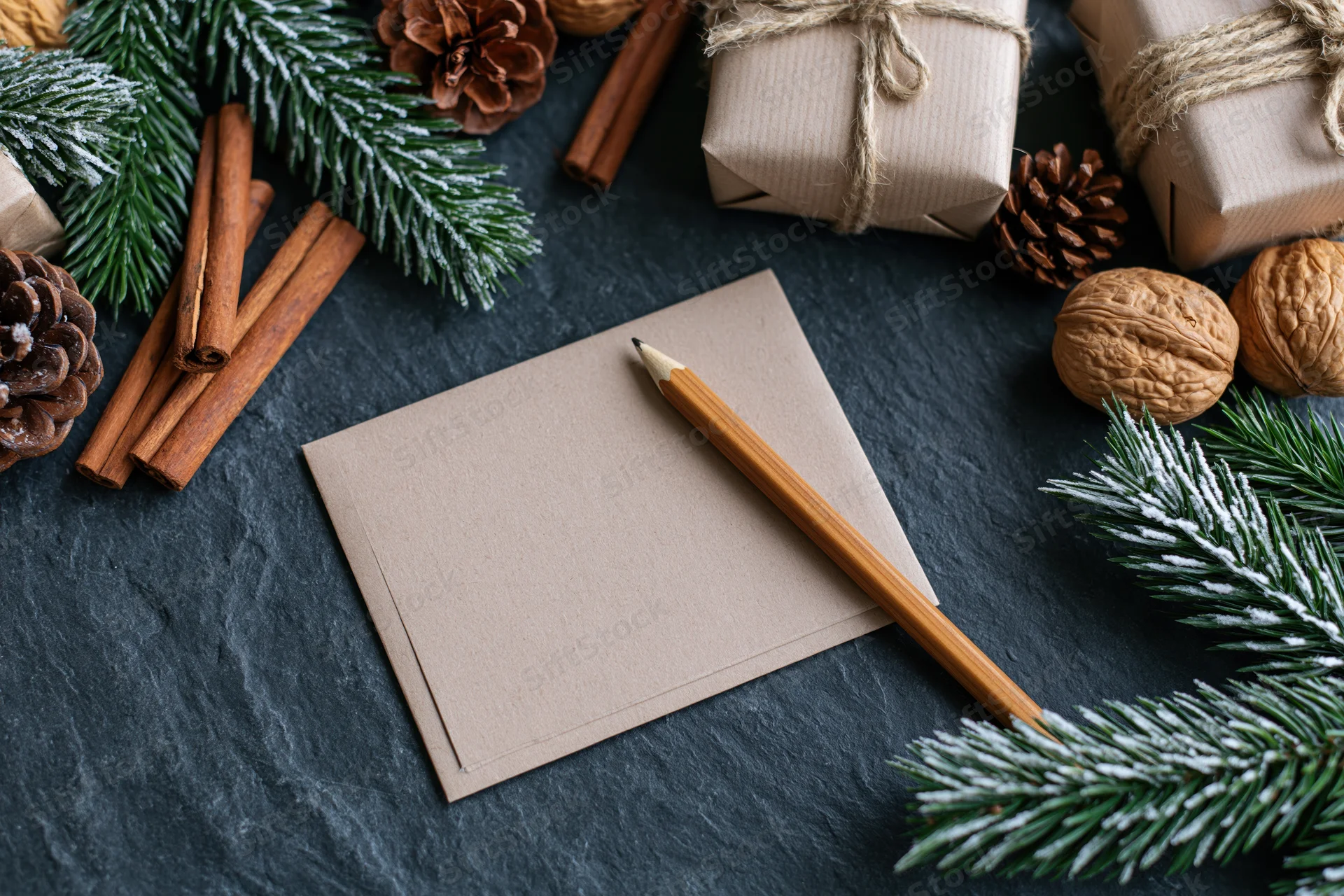 Blank kraft notecard with wooden pencil on dark slate surface surrounded by pine branches, pinecones, cinnamon sticks, walnuts, and wrapped gifts