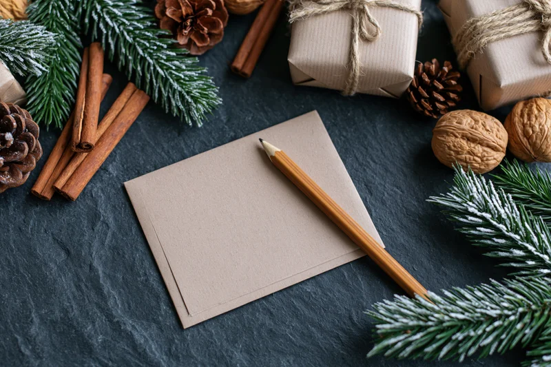 Blank kraft notecard with wooden pencil on dark slate surface surrounded by pine branches, pinecones, cinnamon sticks, walnuts, and wrapped gifts
