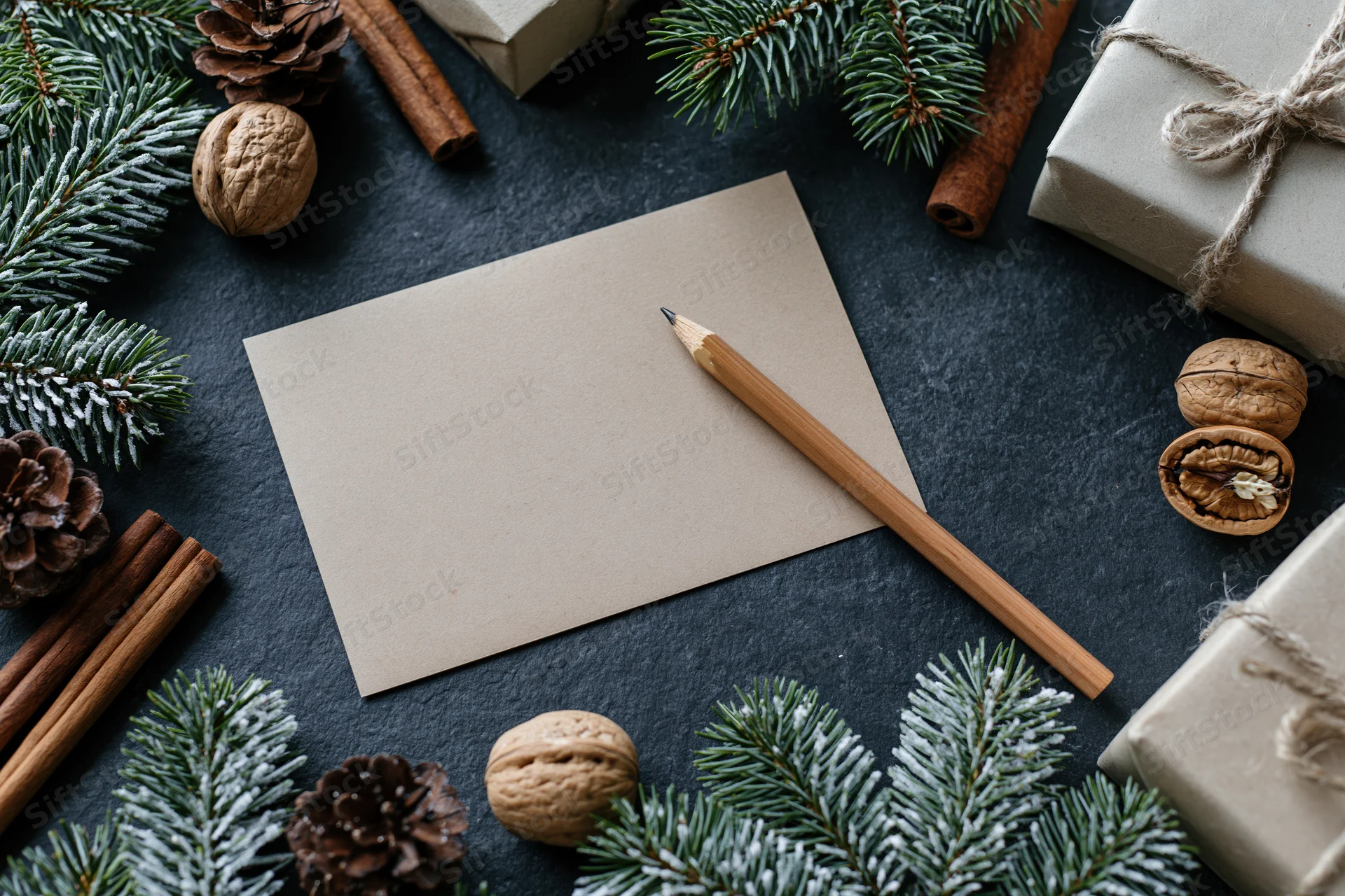 Blank kraft paper notecard with wooden pencil on dark slate surface surrounded by pine branches, pinecones, cinnamon sticks, walnuts, and wrapped gifts