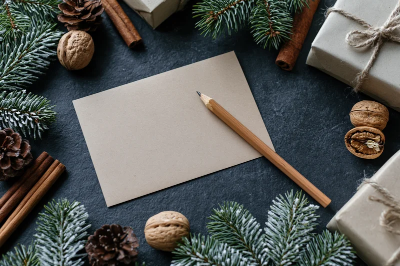 Blank kraft paper notecard with wooden pencil on dark slate surface surrounded by pine branches, pinecones, cinnamon sticks, walnuts, and wrapped gifts