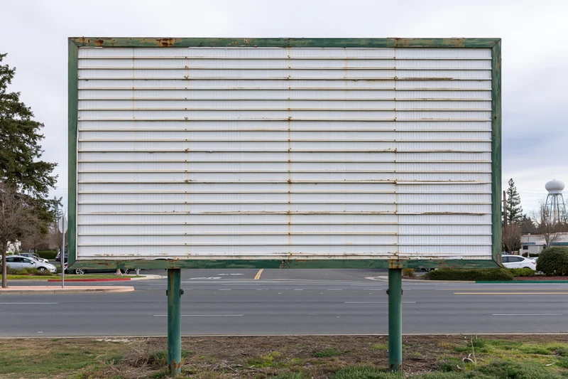 Weathered white billboard with green frame standing beside a road under an overcast sky