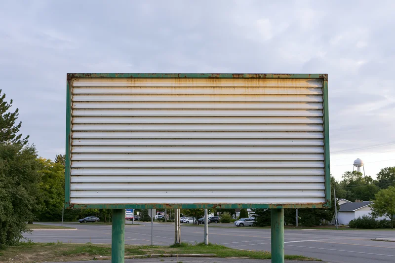 Weathered outdoor billboard with horizontal metal slats mounted on green posts beside a parking lot