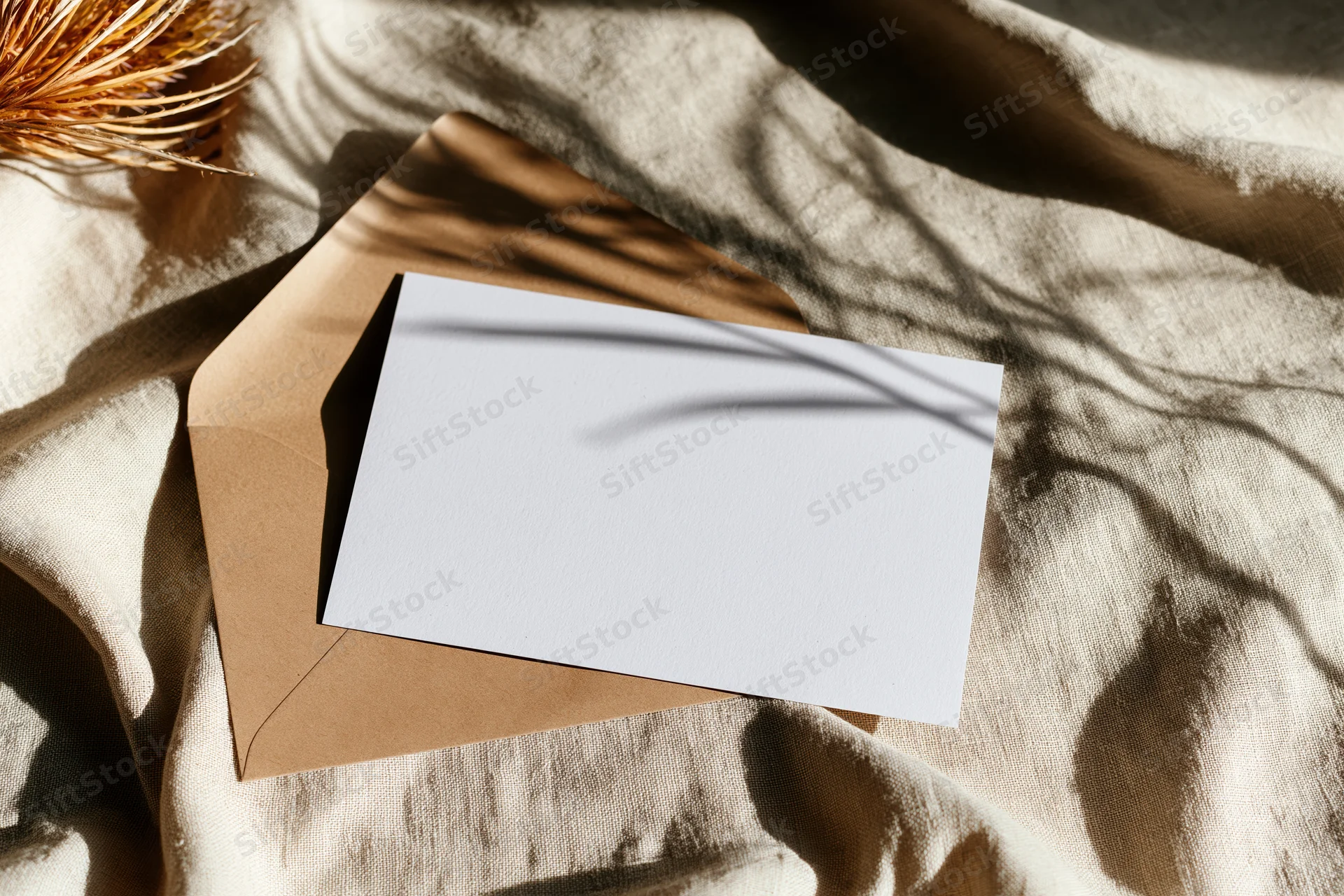 Blank white card on kraft envelope resting on textured fabric with dappled sunlight