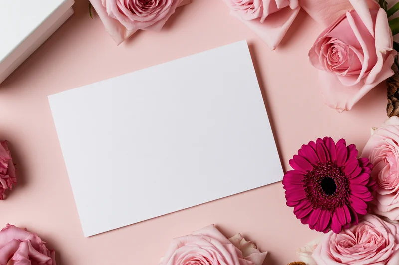 Blank white greeting card on pink surface surrounded by roses and gerbera flowers