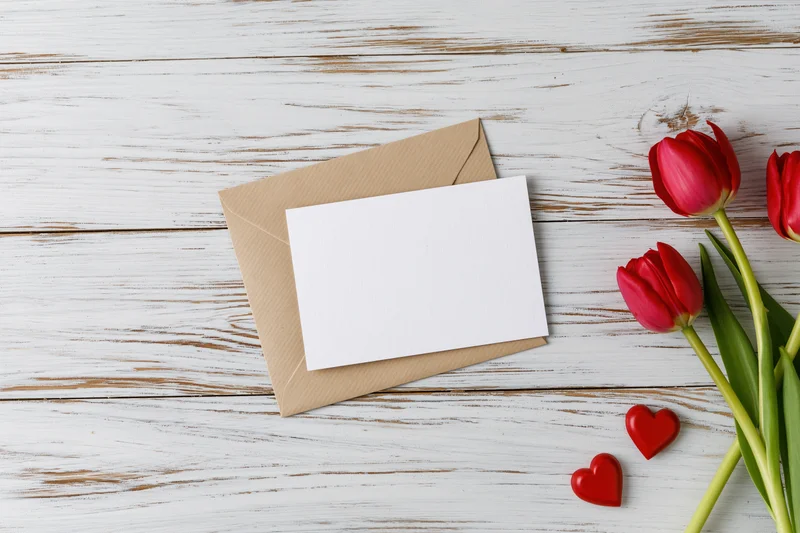 Blank white greeting card on kraft envelope with red tulips and hearts on whitewashed wood surface