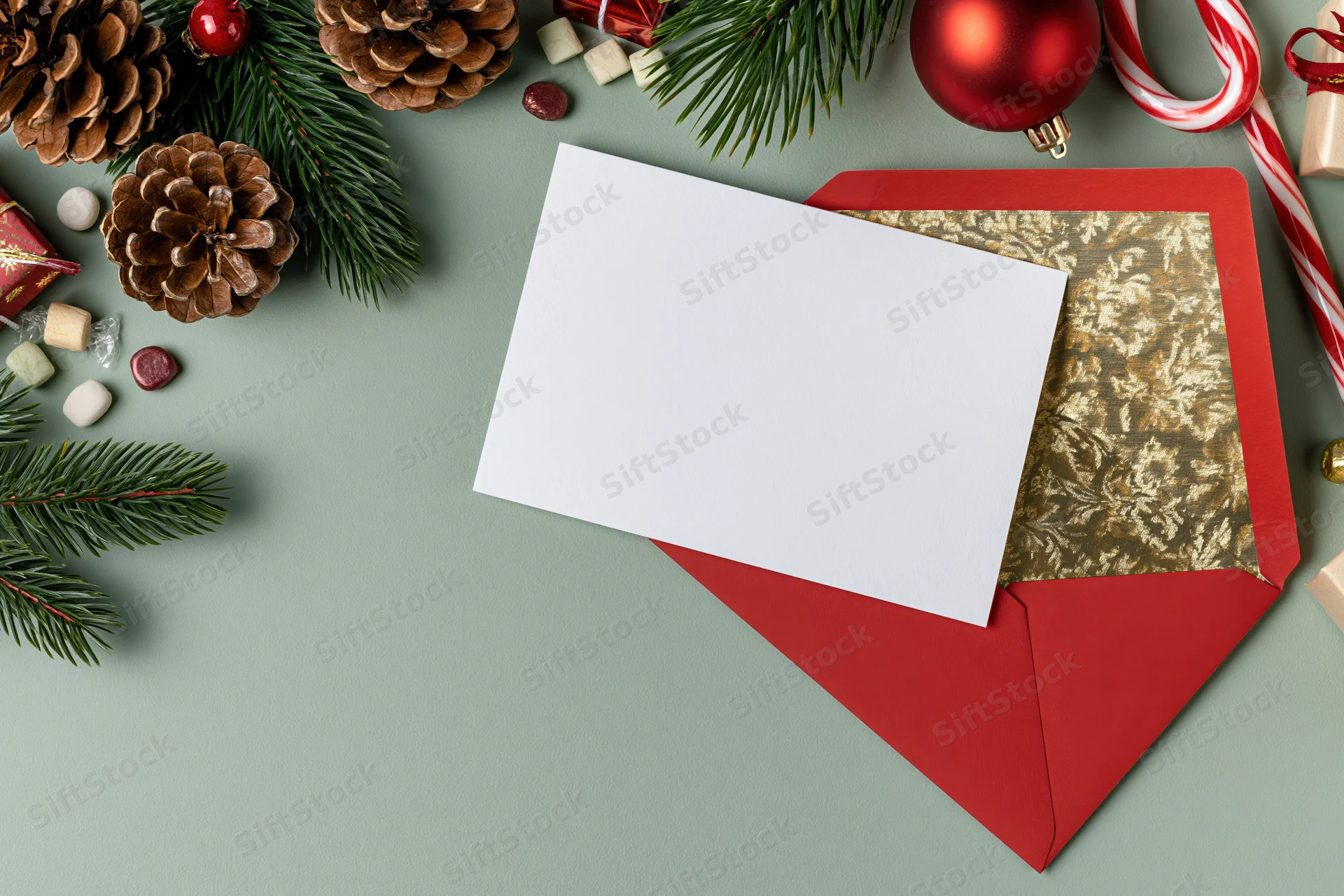 Blank white greeting card on open red envelope with gold foil lining surrounded by christmas decorations on green surface