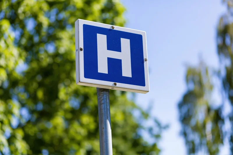 Blue and white hospital directional sign on metal pole outdoors with trees in background