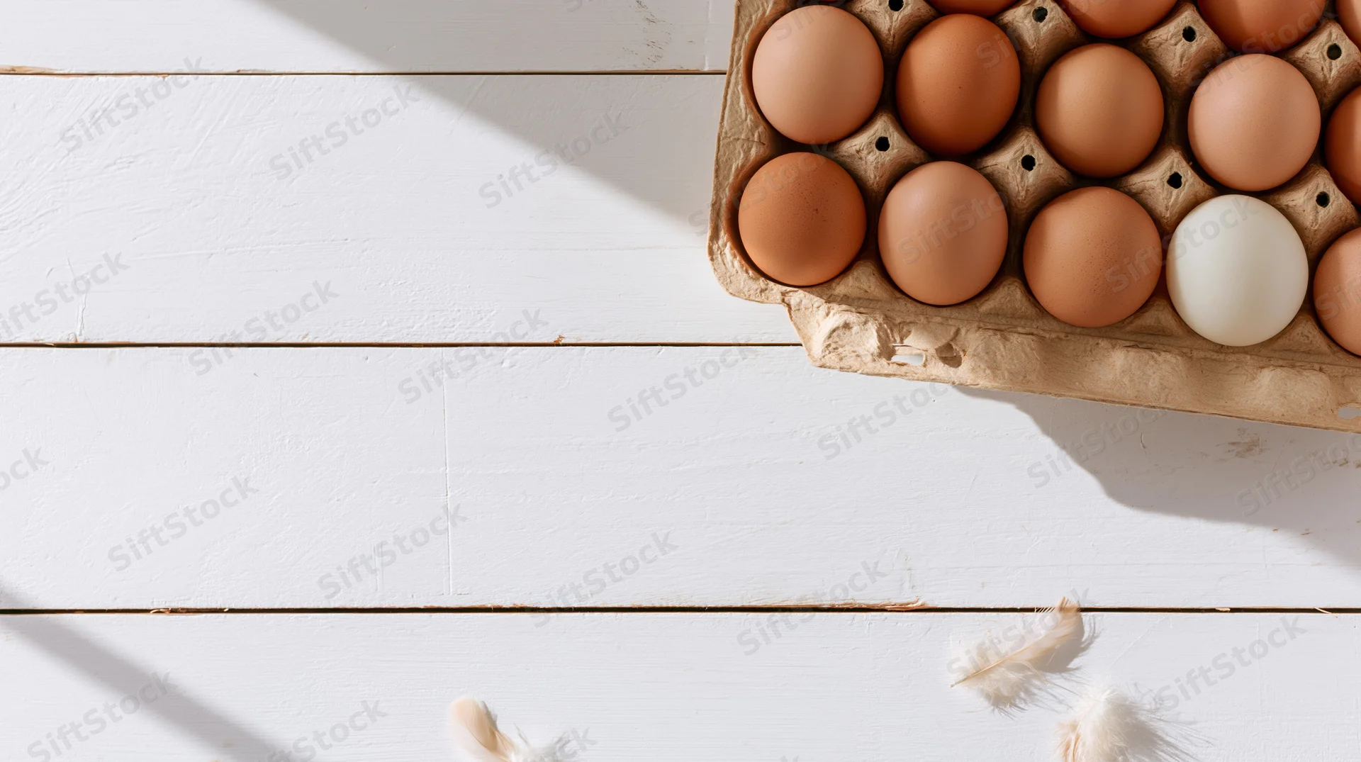 Brown and white chicken eggs in a cardboard carton on a white wooden table with feathers scattered nearby