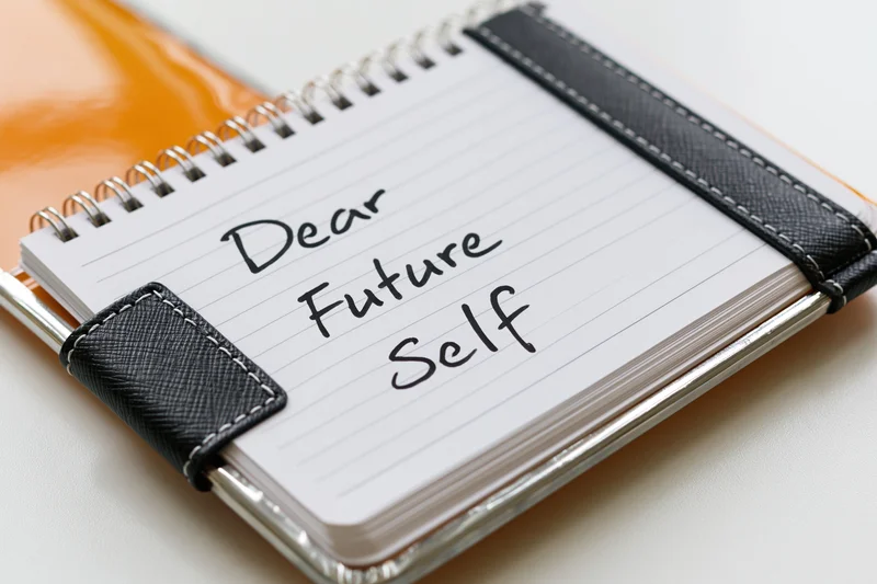 Open spiral notebook with handwritten message "Dear Future Self" on lined paper, secured with black leather strap, resting on orange cover and white surface