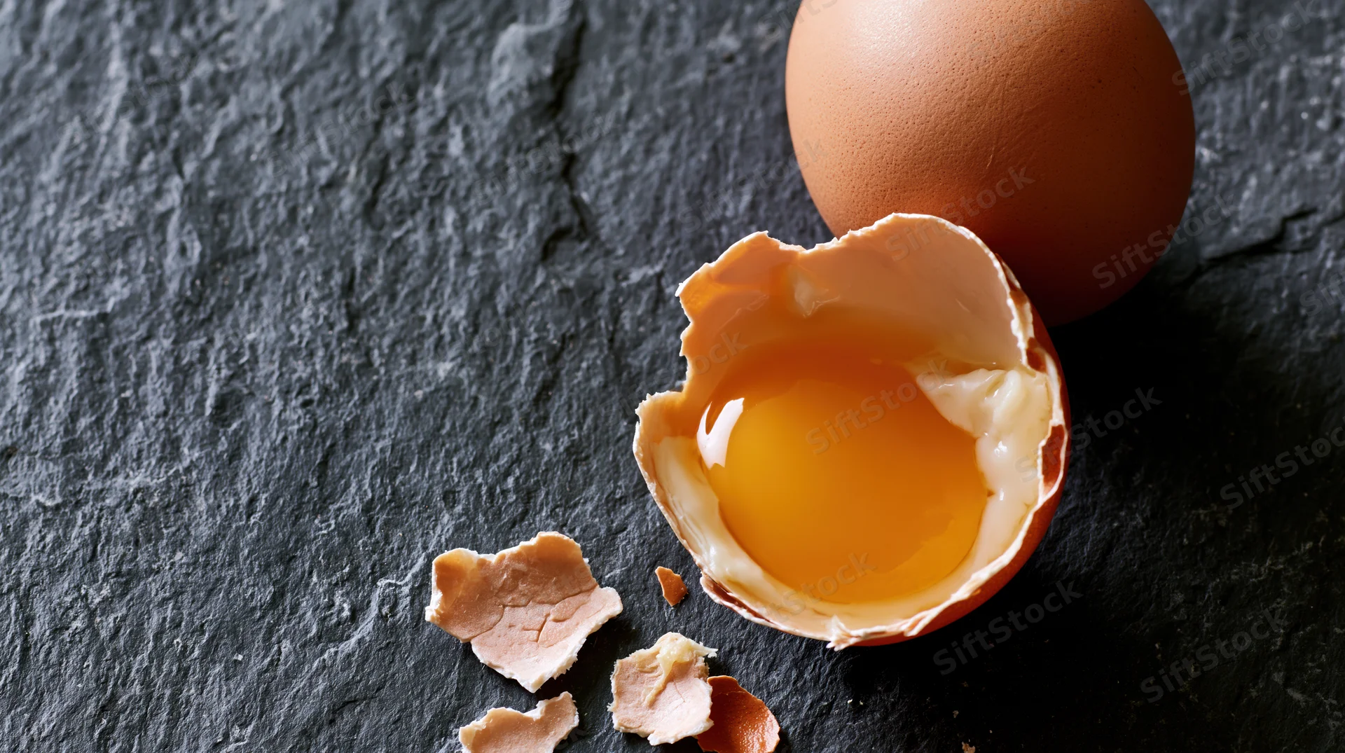 Cracked brown chicken egg with yolk and shell fragments on dark slate stone surface