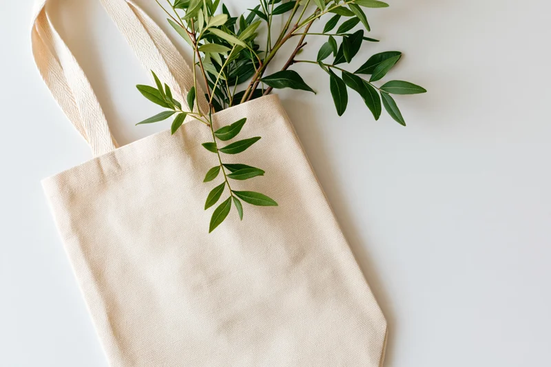Cream canvas tote bag with green leaves spilling out, lying flat on light off-white surface