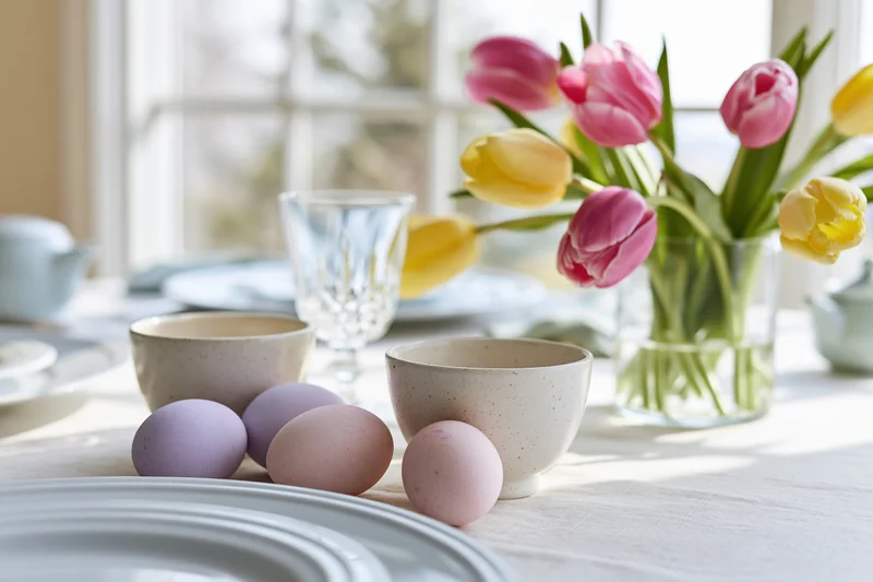 Easter brunch table set with pastel-dyed eggs and ceramic cups near a window with tulips in a vase