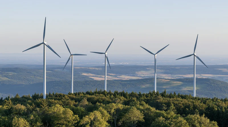 Five wind turbines rising above a forested hilltop in rural countryside