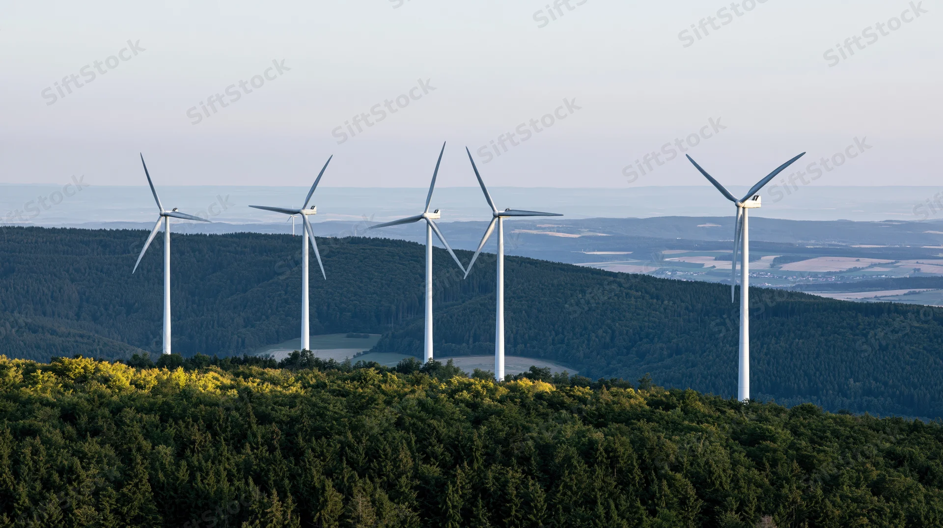 Five wind turbines rising above a forested hilltop in rural countryside
