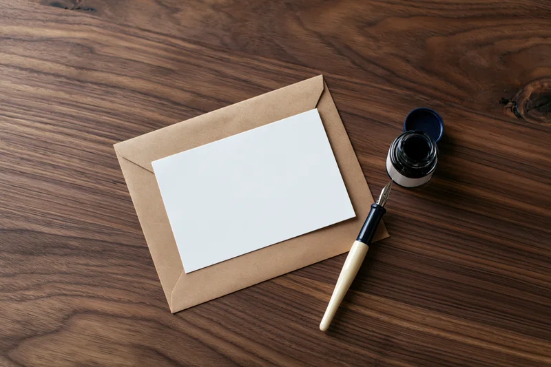Blank white card on kraft envelope with fountain pen and ink bottle on wooden desk