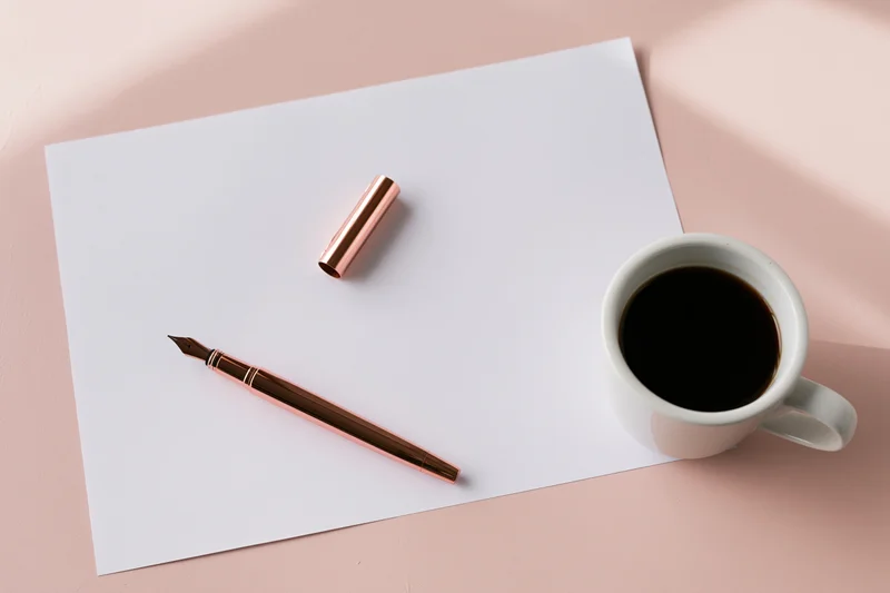 Blank white paper with rose gold fountain pen and coffee cup on pink surface