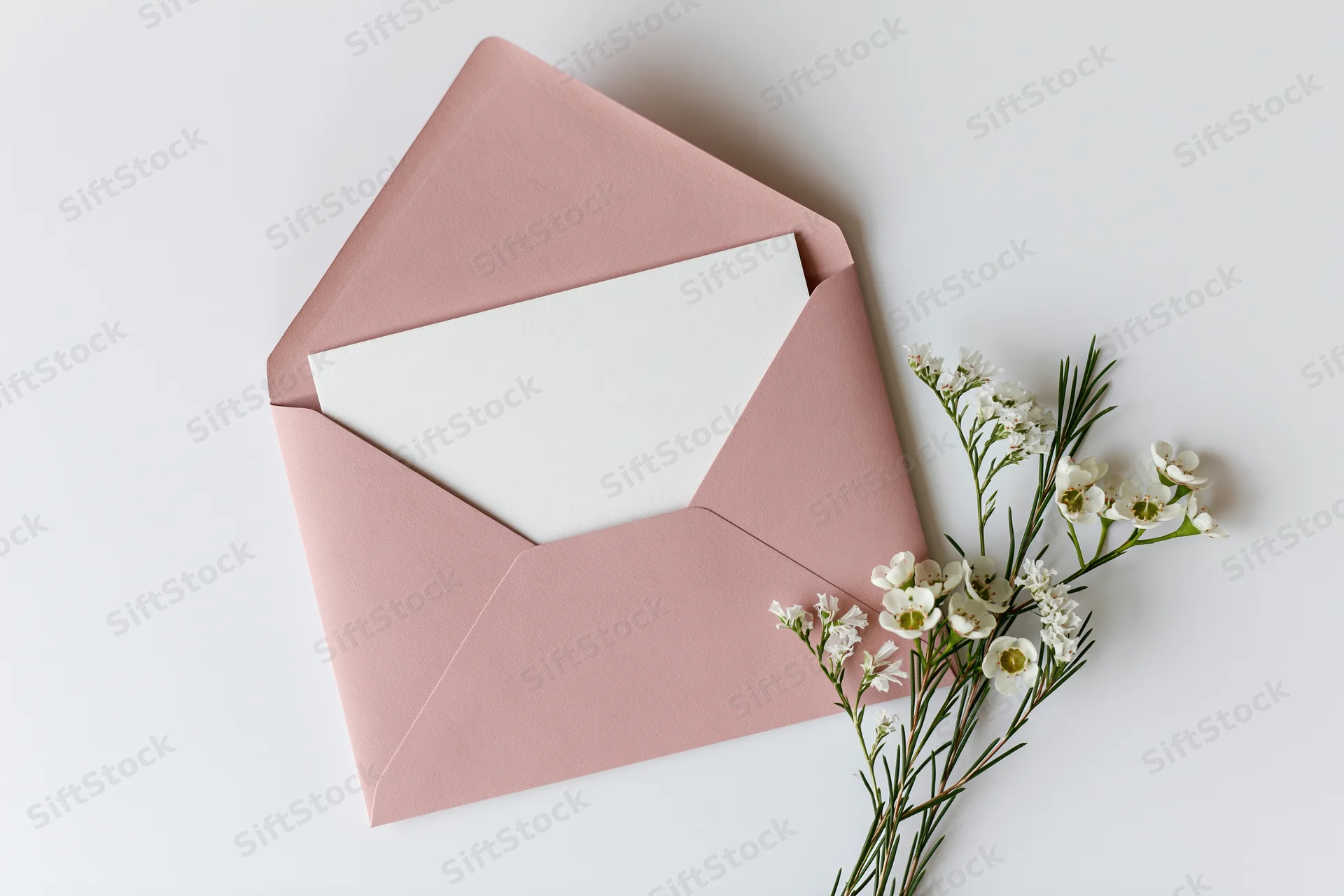 Open dusty pink envelope with blank white paper and white flowers on white surface