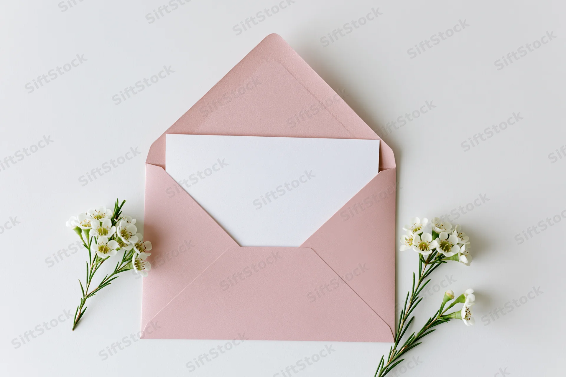 Open dusty pink envelope with blank white card and small white flowers arranged flat on white surface