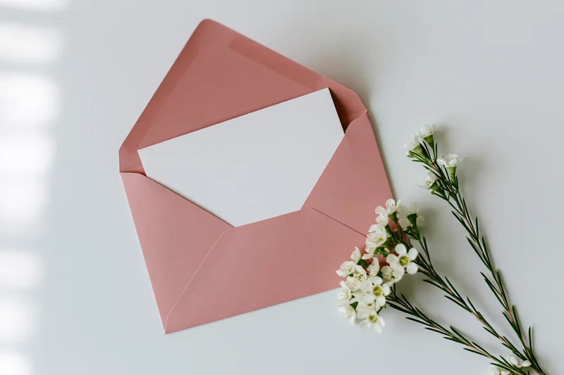 Open dusty pink envelope with blank white paper and white flowers on light surface