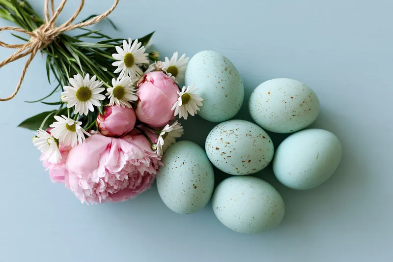 Flat lay of speckled mint easter eggs and a bouquet of pink peonies and white daisies on a light blue surface