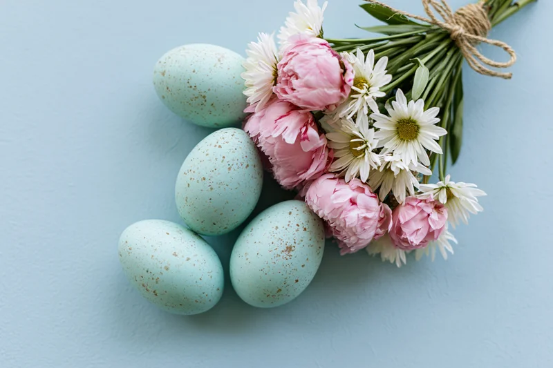 Five speckled mint easter eggs and a bouquet of pink peonies and white daisies on a light blue surface