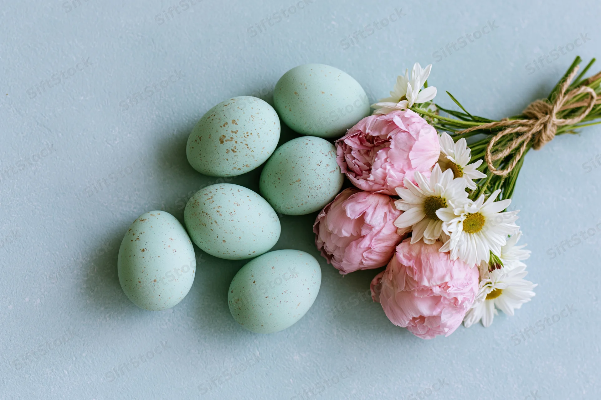 Five pale mint speckled easter eggs and a small bouquet of pink peonies and white daisies arranged on a light blue surface