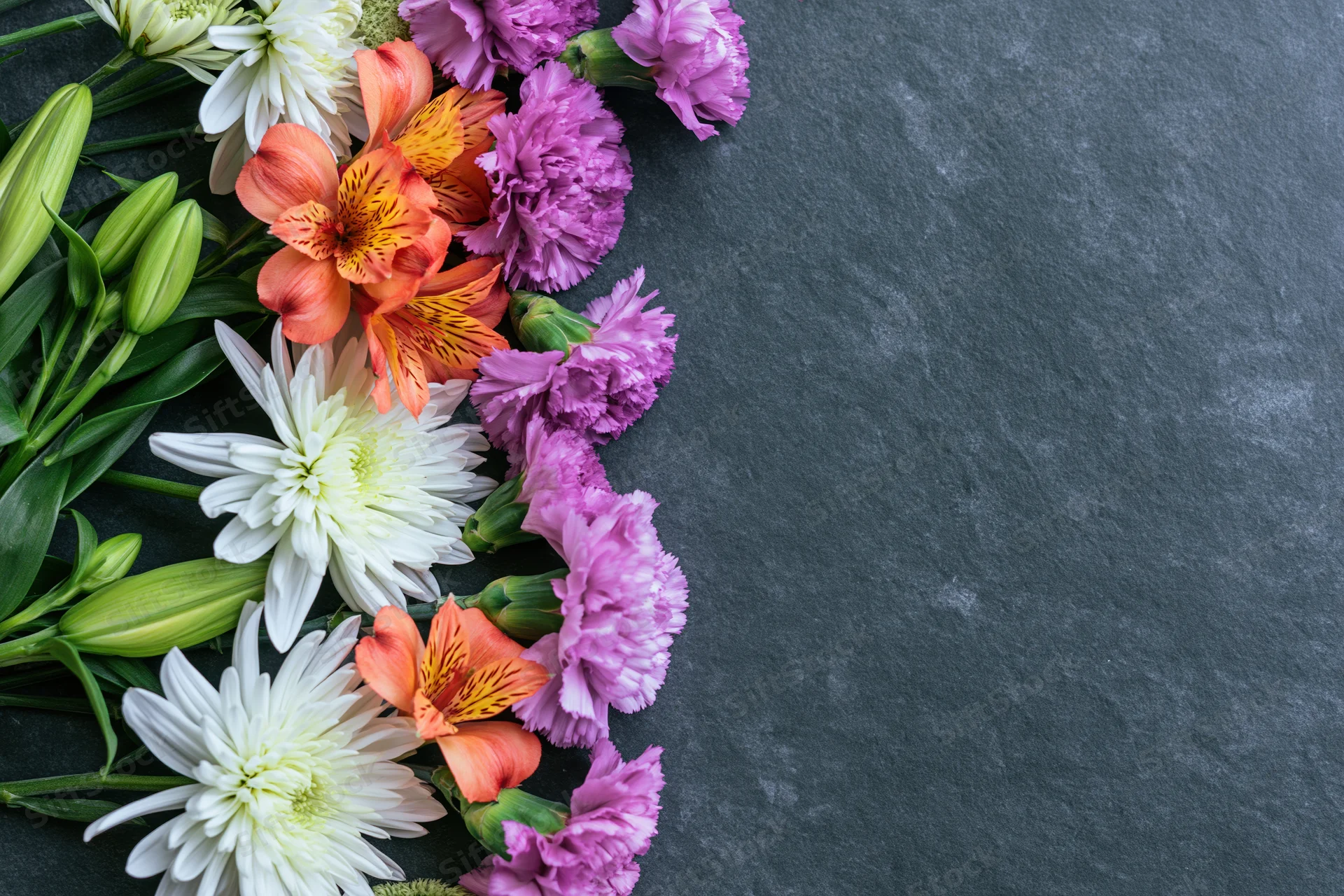 Mixed spring flowers arranged in a flat lay along the left side of a dark slate surface