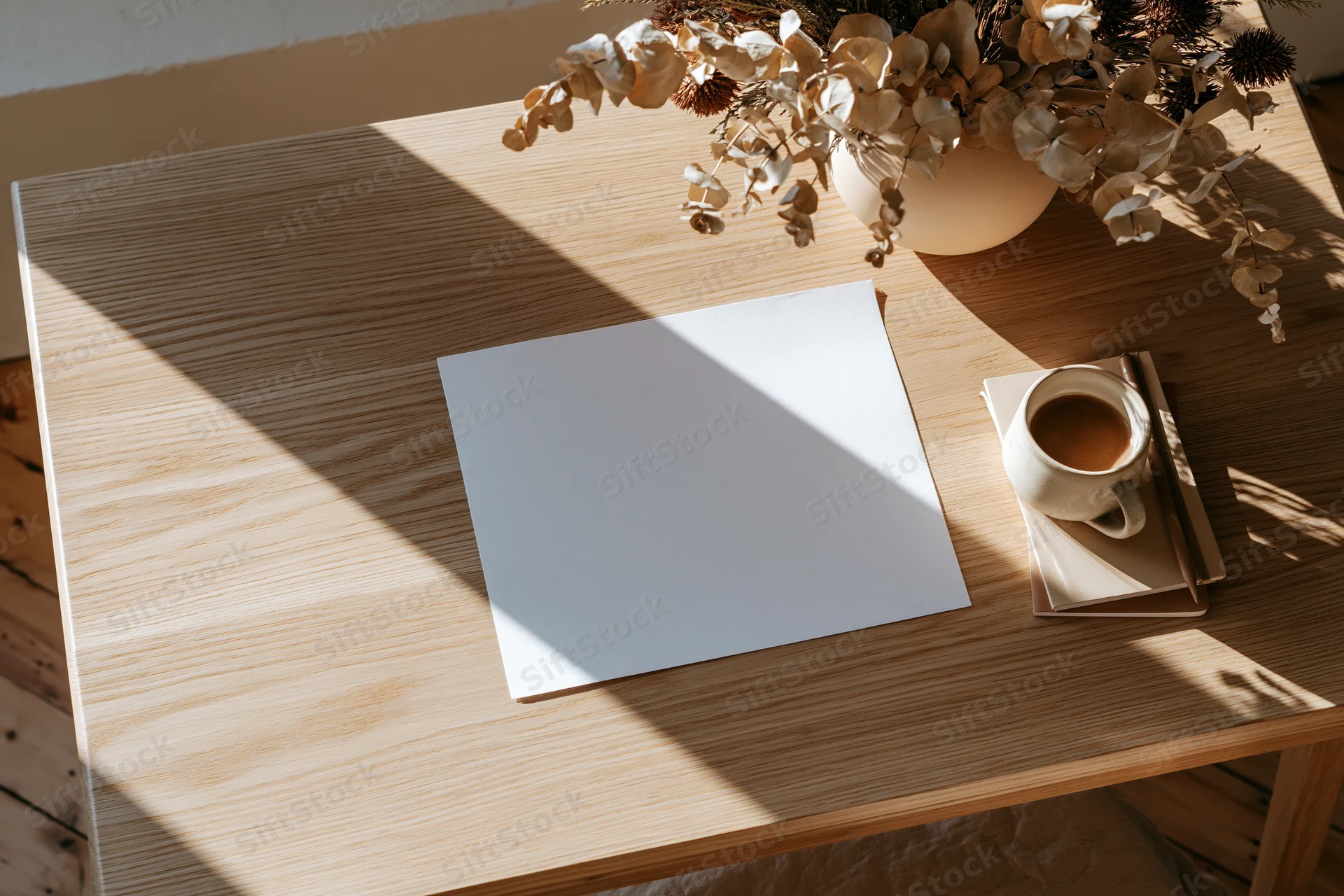 Overhead view of a wooden desk with blank paper, coffee cup, and dried flowers in a vase, lit by sunlight casting shadows