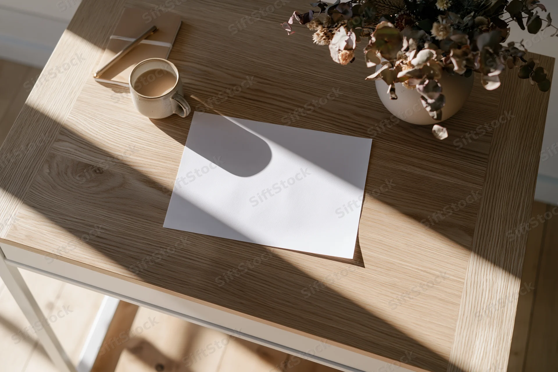 Overhead view of a wooden desk with blank paper, coffee mug, dried flowers, and notebook in natural sunlight