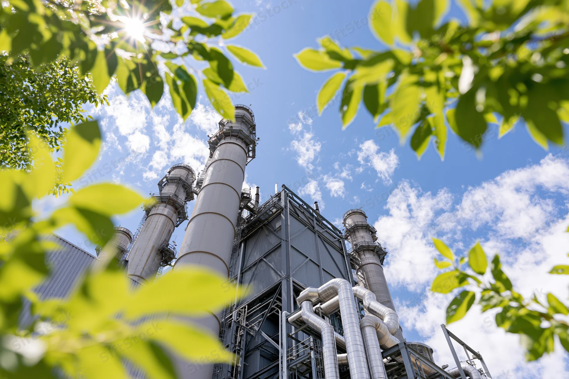 Low-angle view of industrial power plant with tall metal stacks and pipes framed by green leaves against a blue sky with sunlight and clouds