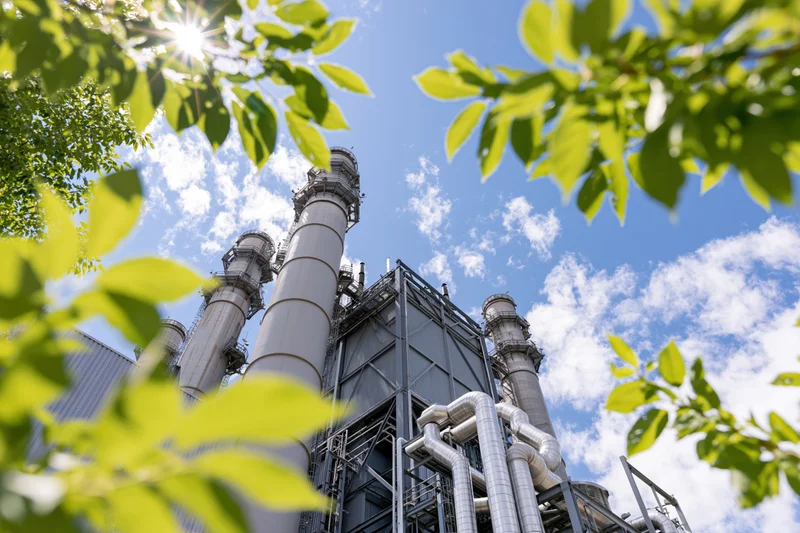 Low-angle view of industrial power plant with tall metal stacks and pipes framed by green leaves against a blue sky with sunlight and clouds