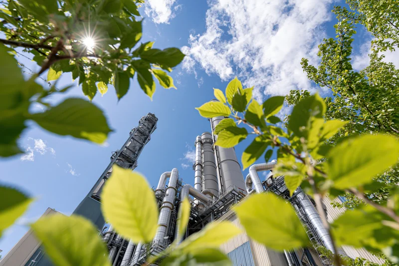 Low-angle view of industrial power plant with tall metal stacks and pipes framed by green leaves against a blue sky with sun and clouds