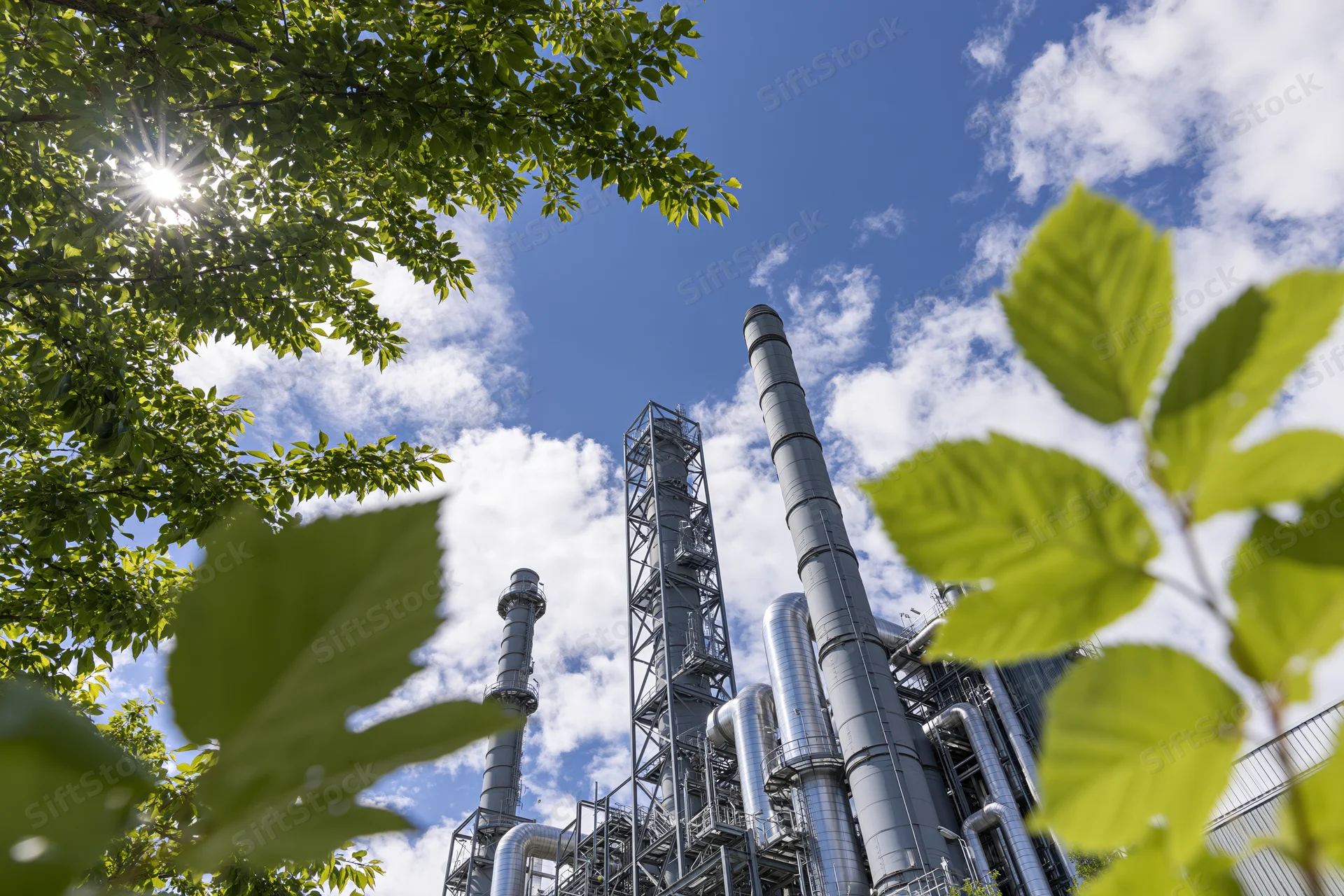 Industrial power plant with tall metal smokestacks and pipes viewed through green tree leaves against a blue sky with clouds