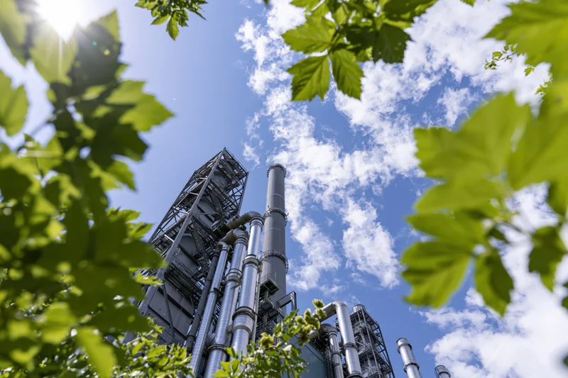 Industrial power plant with tall metal smokestacks and pipes viewed through green leaves against a blue sky
