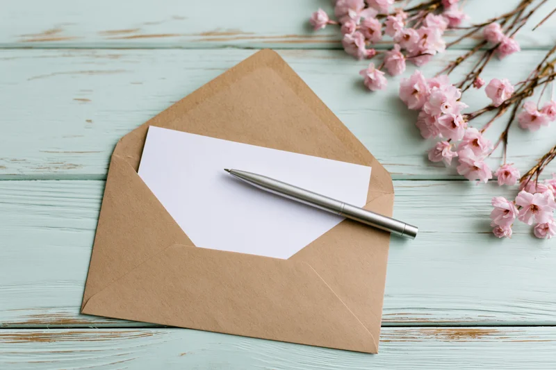 Open kraft envelope with blank white card and silver pen on mint wooden table beside pink cherry blossoms
