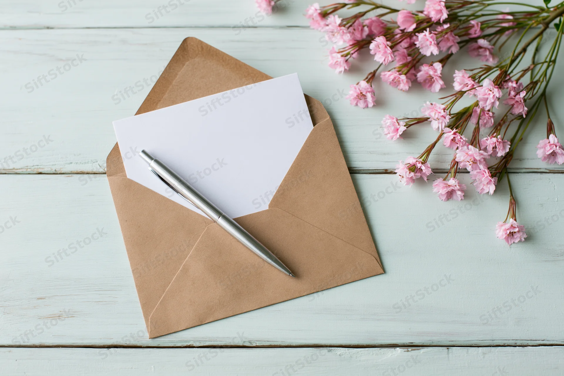 Open kraft envelope with blank white card and silver pen on light blue wooden table beside pink gypsophila flowers