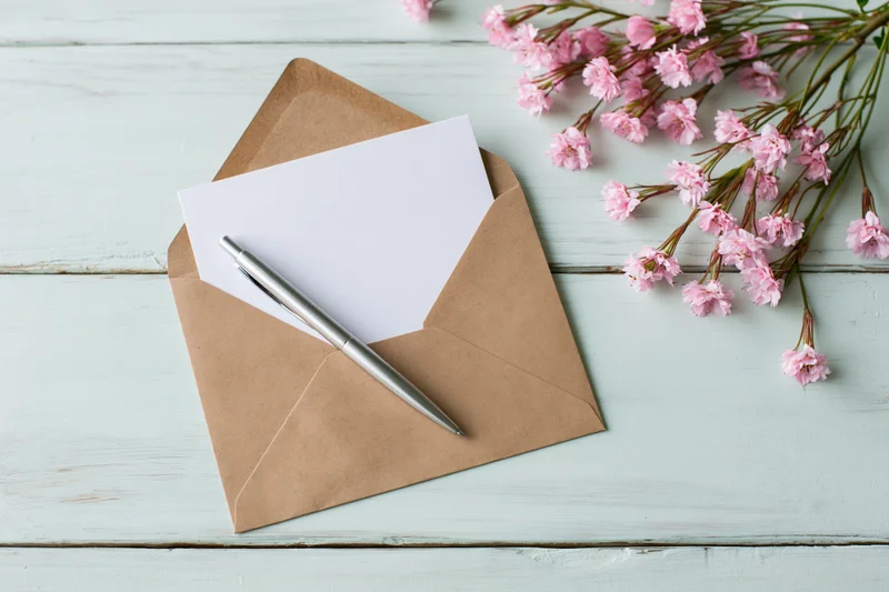 Open kraft envelope with blank white card and silver pen on light blue wooden table beside pink gypsophila flowers