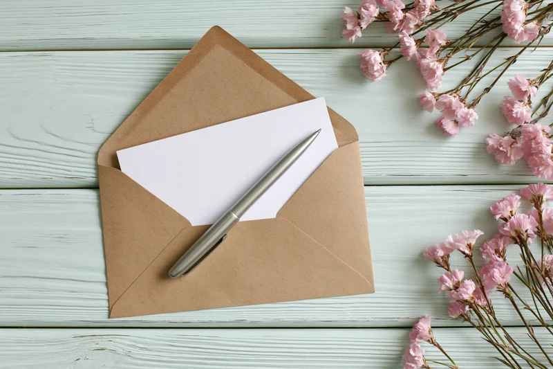 Open kraft envelope with blank white card and silver pen on light blue wooden table beside pink dried carnations