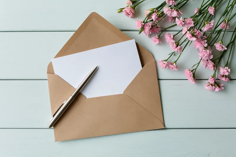 Open kraft envelope with blank white card and silver pen on light blue wooden table beside pink carnation bouquet