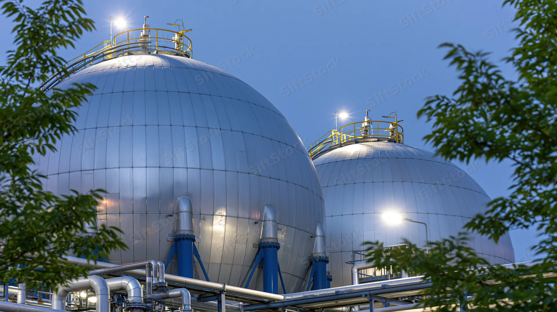 Large spherical steel gas storage tanks at a petrochemical facility with surrounding foliage and evening sky