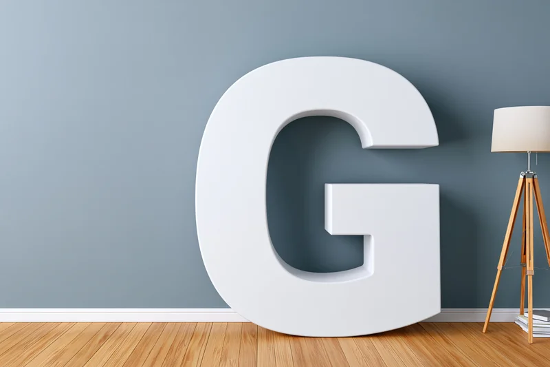 Large white three-dimensional letter g leaning against a gray wall in a minimalist room with wooden floor and tripod lamp