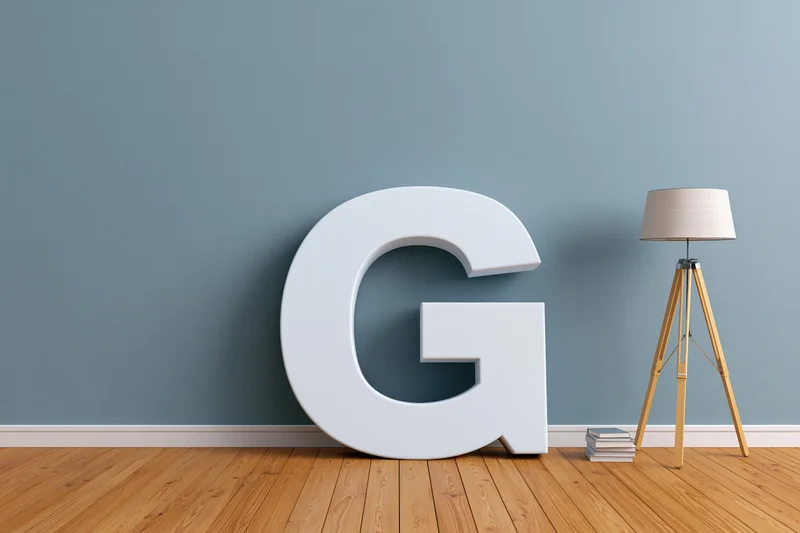 Large white three-dimensional letter g leaning against a blue wall in a minimalist room with wooden floor, tripod lamp and stack of books