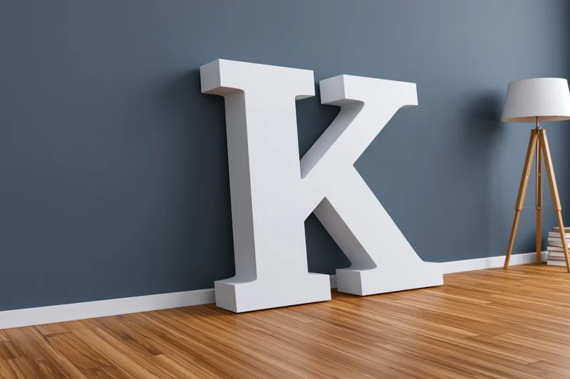 Large white three-dimensional letter k leaning against a dark gray wall in a minimalist room with wooden flooring and a tripod lamp