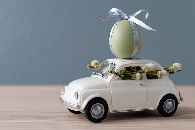 Miniature white vintage toy car carrying a green easter egg with ribbon and willow branches on wooden surface