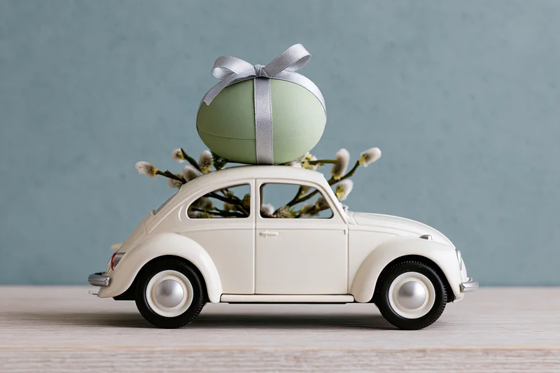 Miniature white vintage toy car carrying a green easter egg with ribbon and willow branches on wooden surface