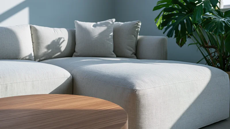 Light grey linen sectional sofa in a modern living room corner with a round wooden coffee table and a monstera plant