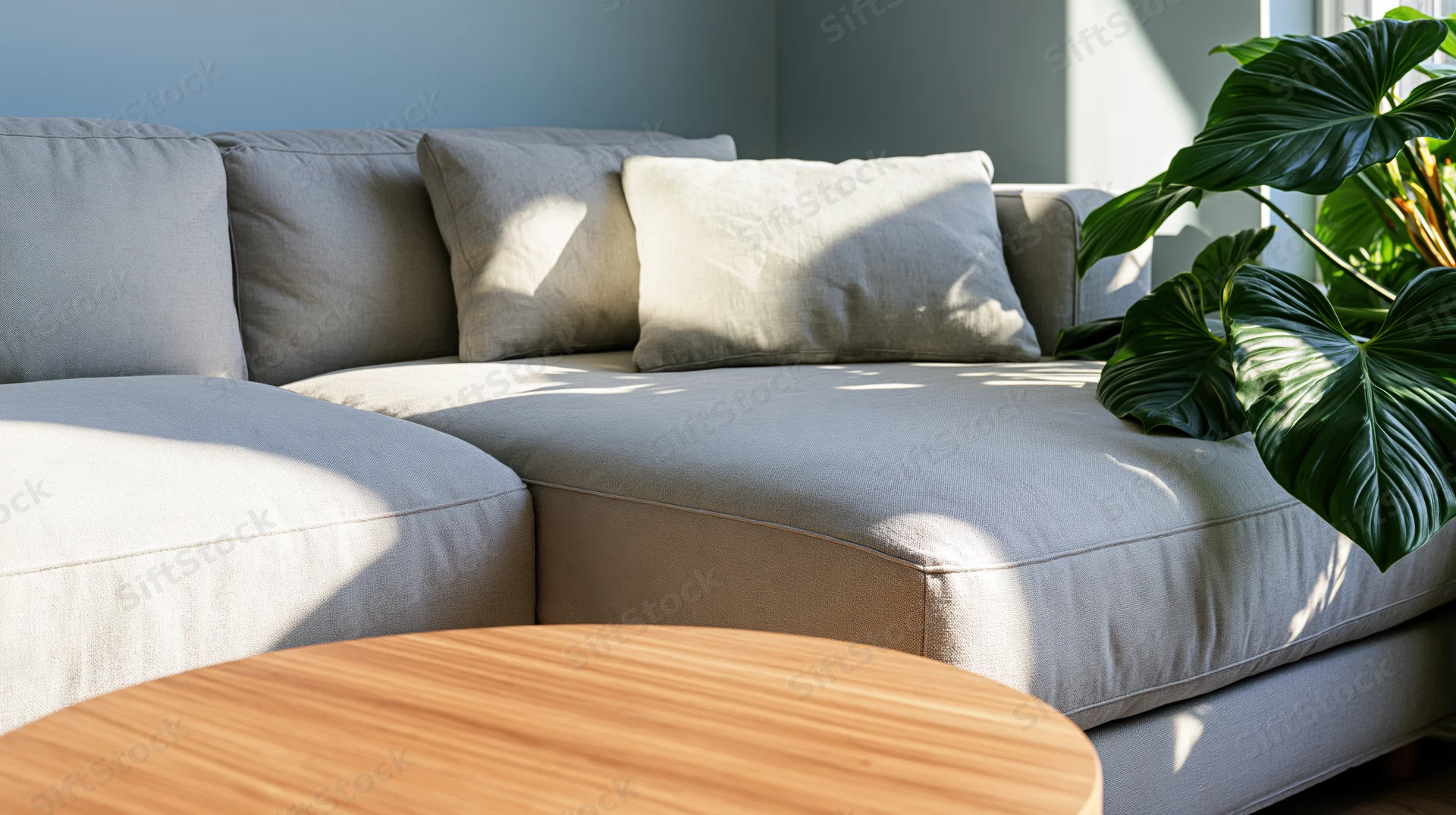 Light grey linen sectional sofa with cushions and a leafy plant in a modern living room corner, sunlight casting shadows on the fabric and a round wooden coffee table in front